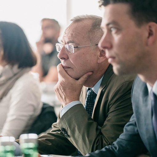 Älterer Mann mit Brille sitzt aufmerksam mit der Hand am Kinn in einem Seminarraum; neben ihm ein junger Mann im Anzug, weitere Teilnehmende im Hintergrund.
