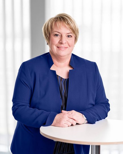 Porträt einer Frau mit kurzem, blondem Haar. Sie trägt ein königsblaues Jackett über einer gemusterten Bluse. Sie steht an einem runden Tisch, hat die Hände locker gefaltet und blickt freundlich in die Kamera. Der Hintergrund ist hell und modern.