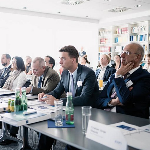 „Gruppe von Teilnehmenden sitzt aufmerksam in einem hellen Seminarraum an einem langen Tisch; auf dem Tisch stehen Wasserflaschen, Gläser und Namensschilder, im Hintergrund ein Regal mit Fachbüchern.