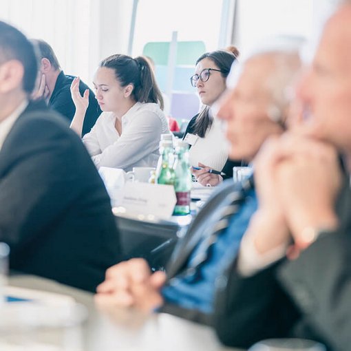 Seminarteilnehmende unterschiedlichen Alters sitzen nebeneinander an einem Tisch und hören aufmerksam zu; im Hintergrund sind Getränke und Notizmaterial zu sehen.