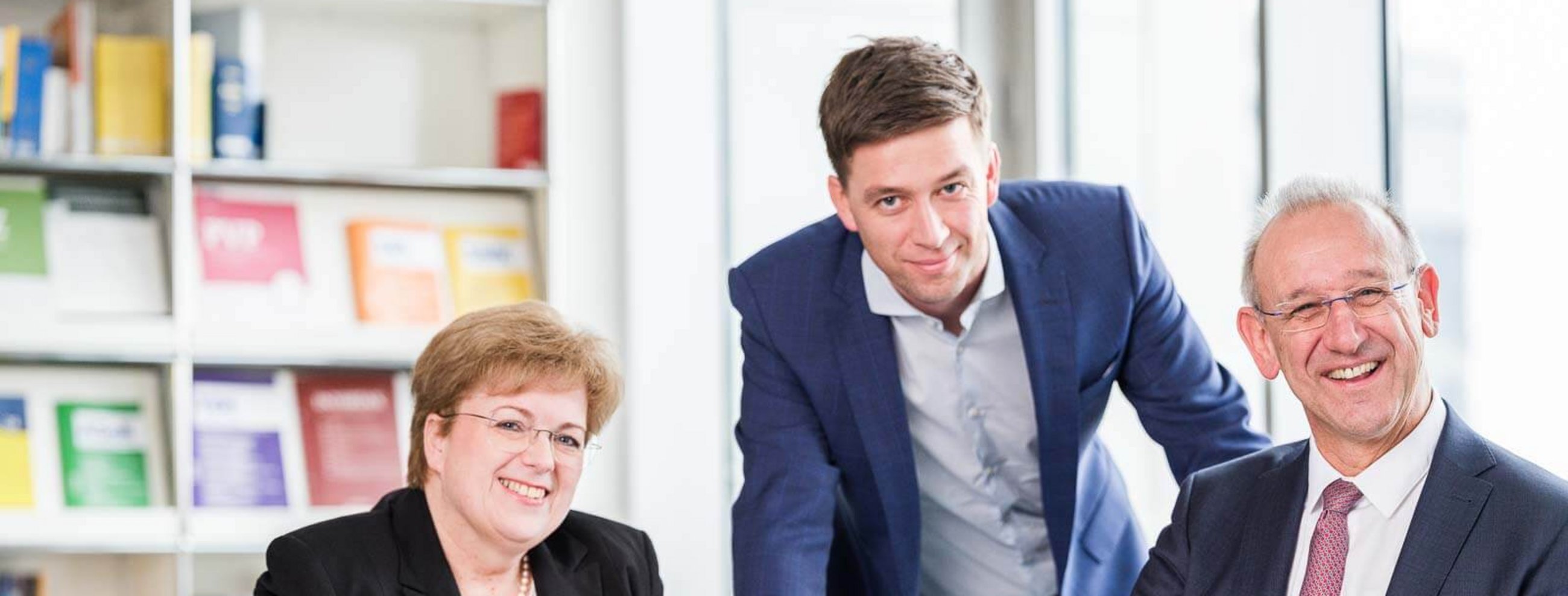 Zwei Männer und eine Frau in Businesskleidung sitzen und stehen an einem Konferenztisch in einem modernen Büro mit Bücherregal im Hintergrund. Alle drei lächeln freundlich in die Kamera.
