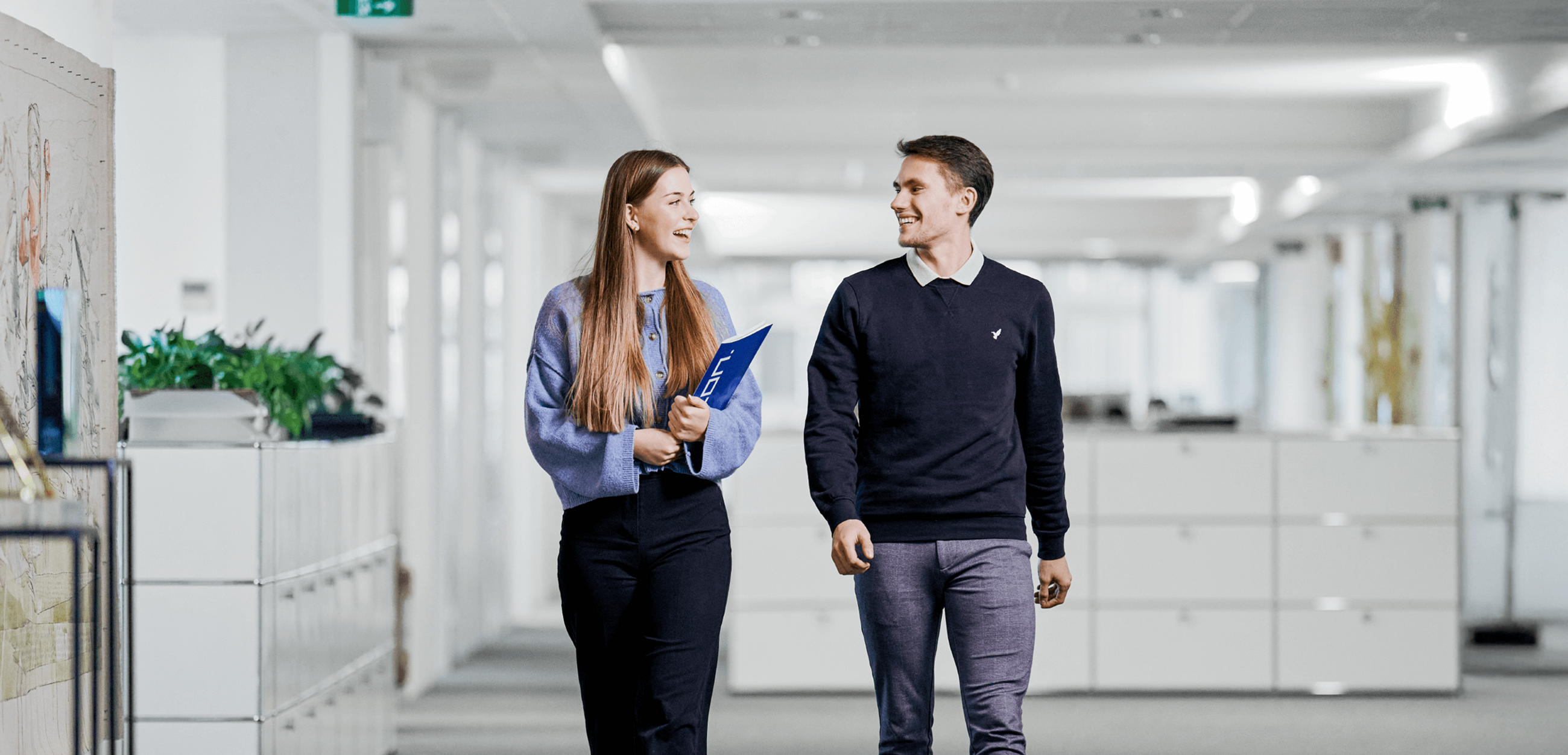 Junge Frau mit Klemmbrett und junger Mann im Gespräch, gemeinsam gehend durch einen modernen, hellen Büro Flur.