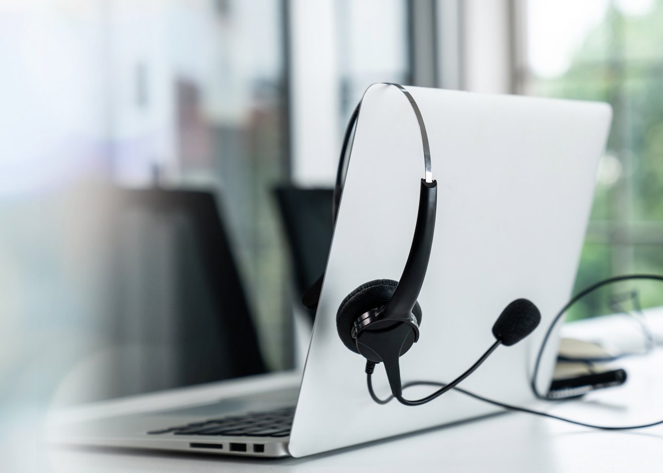 Das Bild zeigt einen weißen Laptop mit einem angeschlossenen Headset. Der Hintergrund ist unscharf und zeigt eine moderne Büroumgebung mit Fenstern.