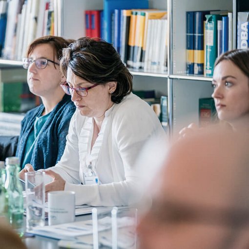 Drei Frauen sitzen aufmerksam nebeneinander an einem Tisch in einem Seminarraum mit Bücherregalen im Hintergrund; im Vordergrund ist verschwommen ein weiterer Kopf zu sehen.