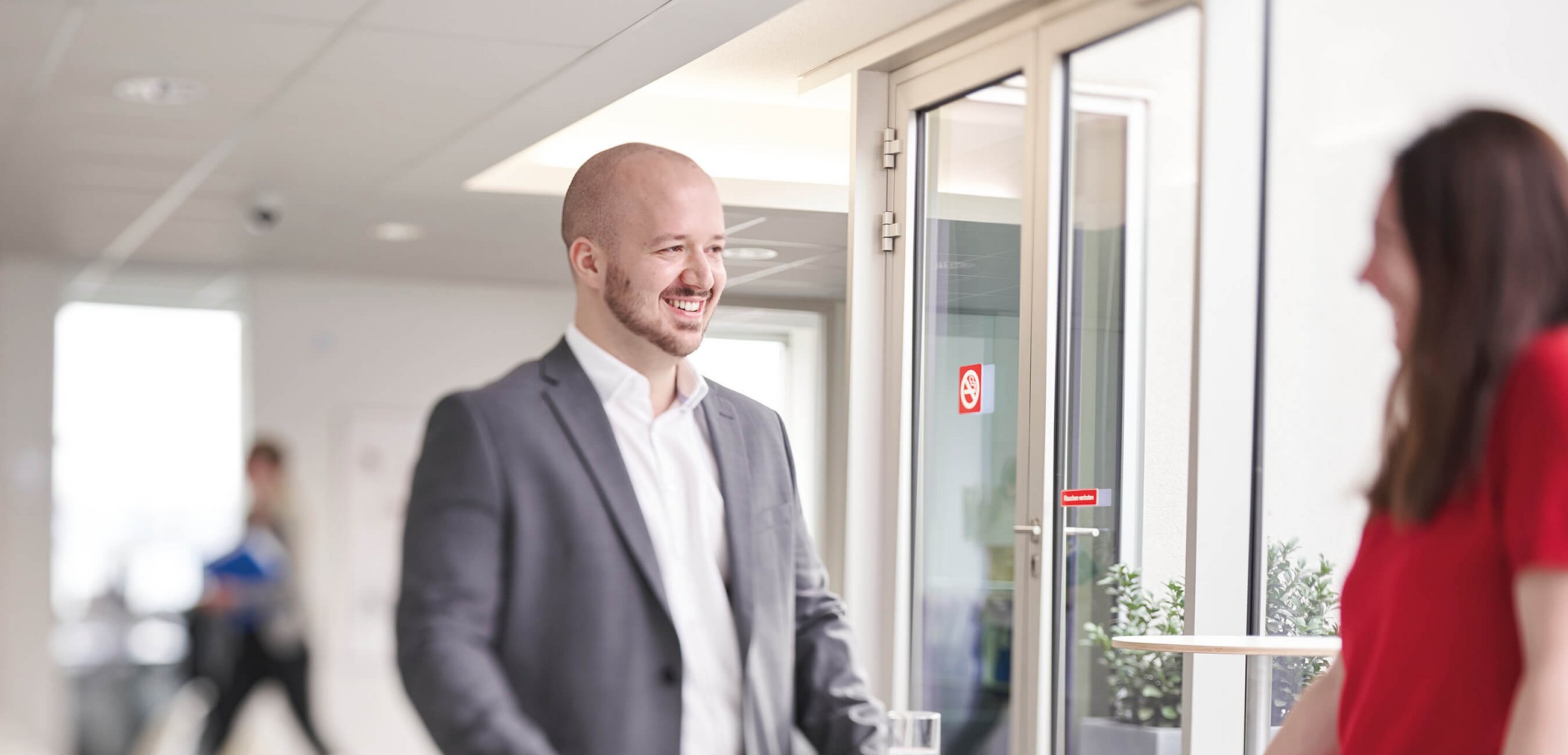 Lächelnder Mann im grauen Anzug steht in einem hellen Büroflur im Gespräch mit einer Frau in rotem Oberteil; im Hintergrund Glastüren und weitere Personen.