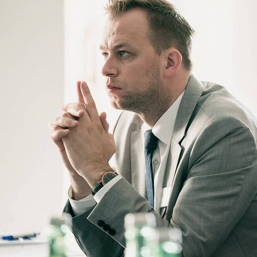 Junger Mann im grauen Anzug sitzt konzentriert mit gefalteten Händen an einem Seminartisch; im Vordergrund unscharf grüne Glasflaschen.