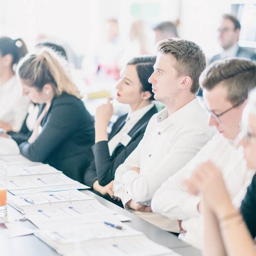 Gruppe junger Menschen sitzt nebeneinander an einem Tisch in einem hellen Seminarraum und folgt aufmerksam dem Geschehen; auf dem Tisch liegen Unterlagen und Getränke.