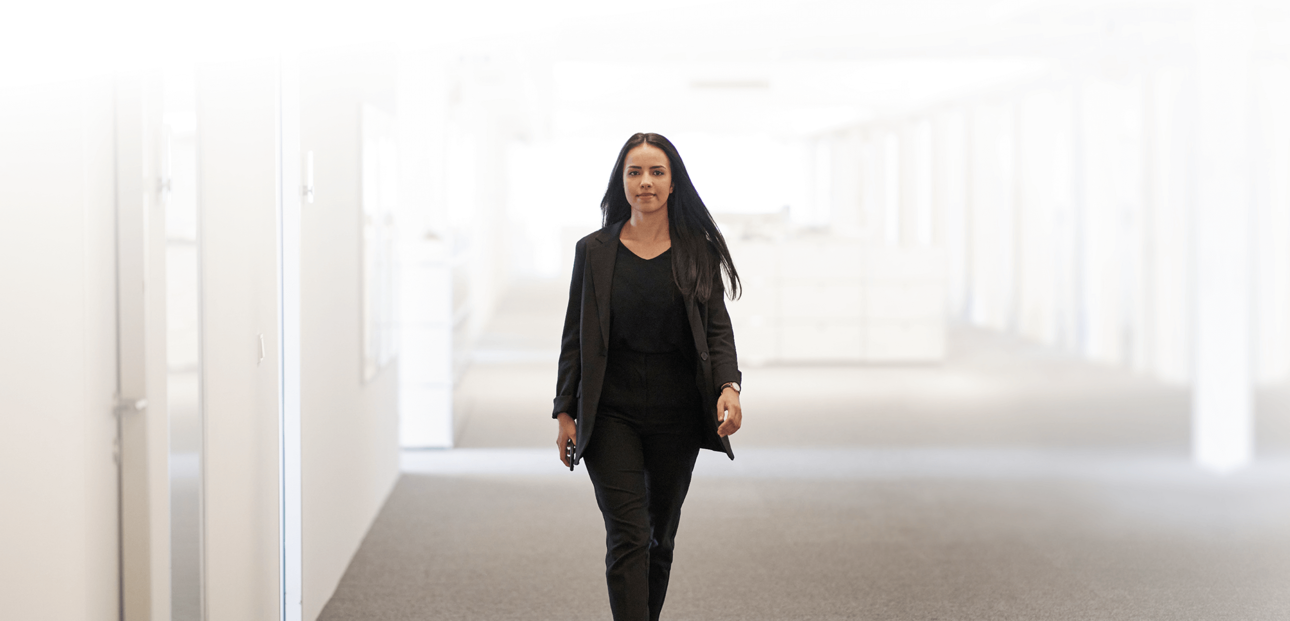 Junge Frau in dunkler Businesskleidung geht selbstbewusst durch einen hellen, modernen Büro Flur.