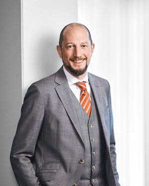 Mann mit kurz rasiertem Haar und Bart steht leicht seitlich vor einem hellen Hintergrund mit Vorhängen. Er trägt einen grauen, dreiteiligen Anzug mit weißem Hemd und orange-gestreifter Krawatte.